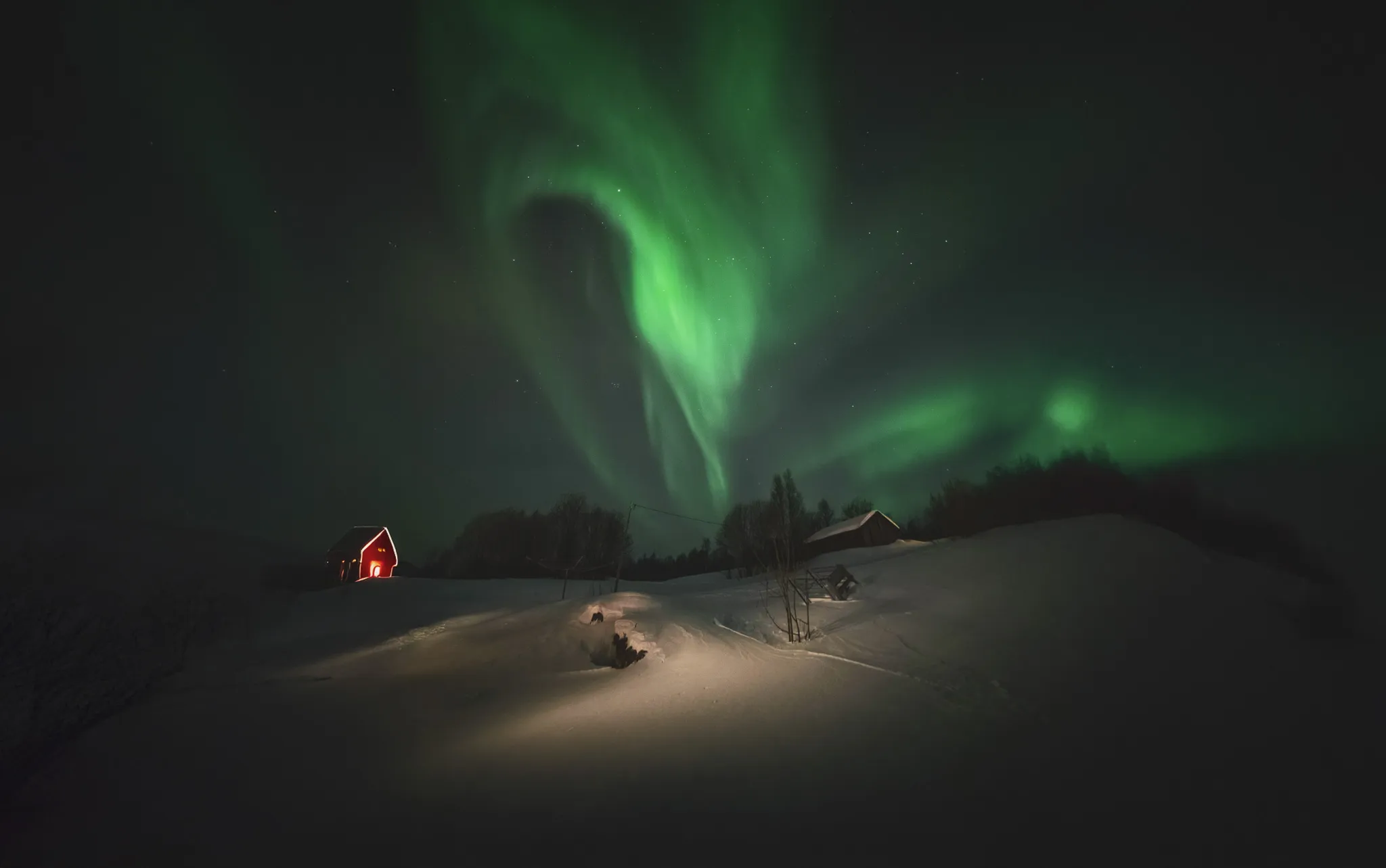 A night landscape illuminated by a brilliant green aurora borealis, unfolding over a snow-covered field. On the left, a small red chalet emits a warm glow, while on the right, a white cottage is illuminated by the light of the aurora borealis.