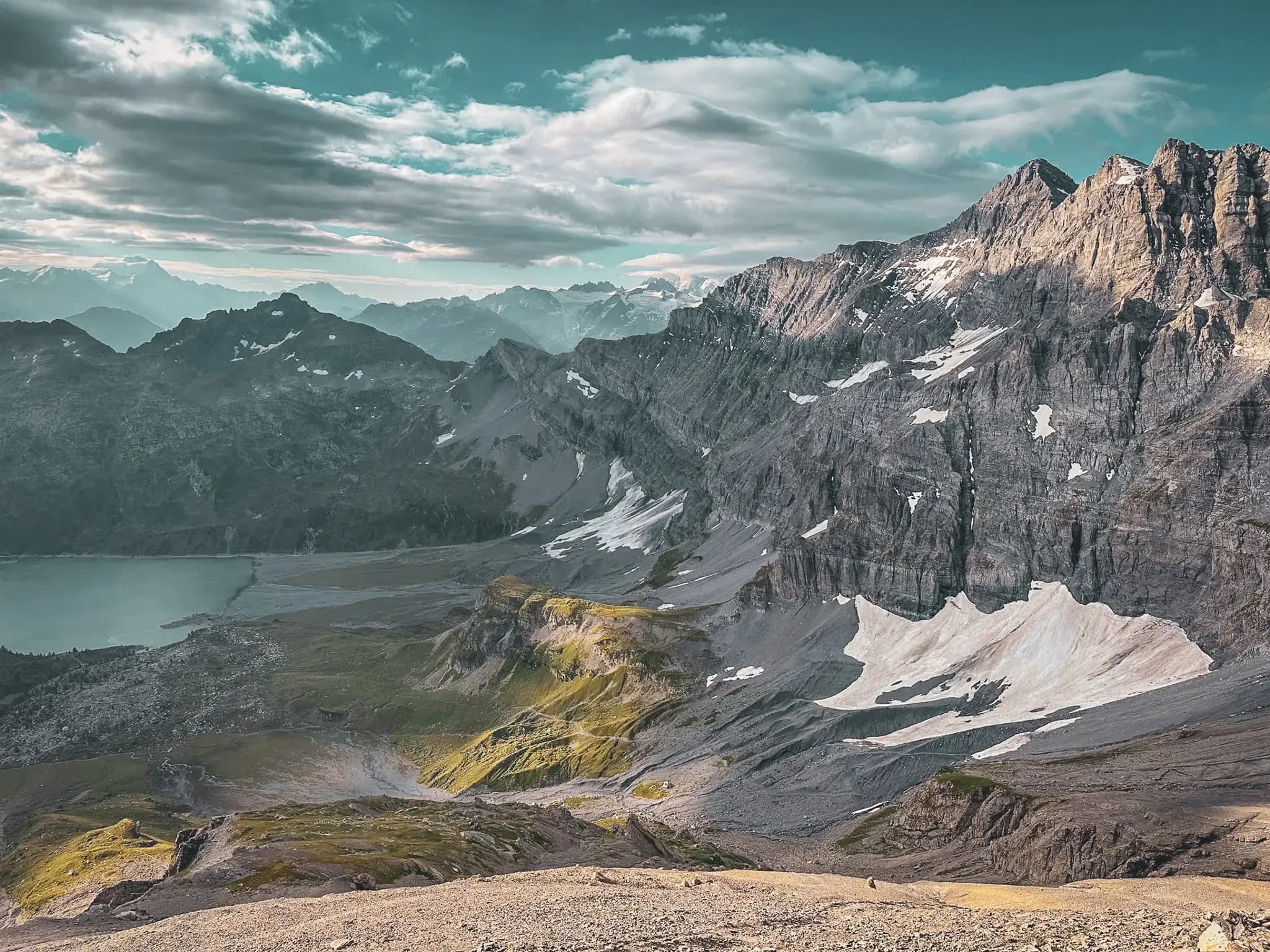 A majestic mountain landscape, with steep peaks and rocky ridges. Persistent snow can be seen on some of the slopes, while below lies a lake with calm waters reflecting the sun's rays.