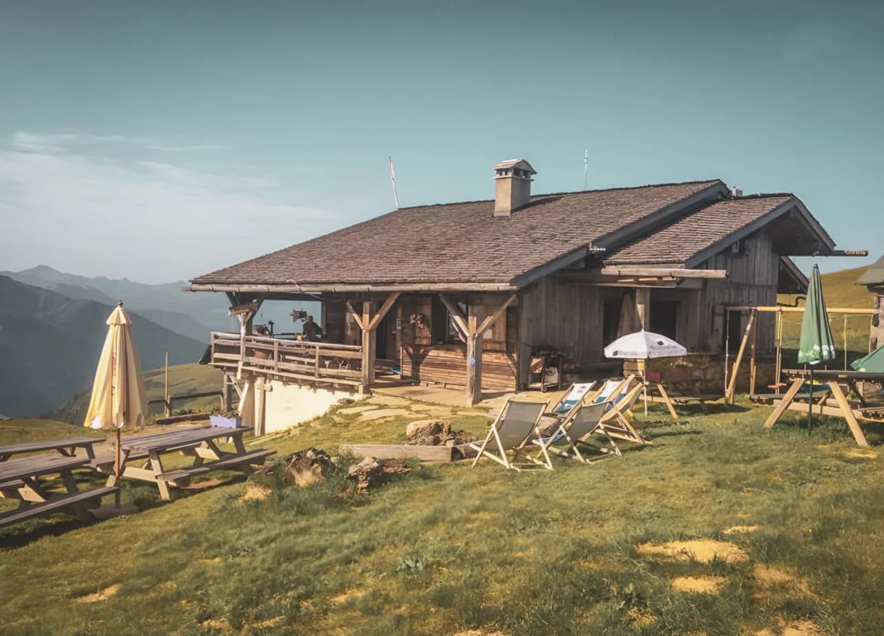 Refuge en bois sur un alpage, entouré de montagnes, avec chaises longues et parasols au soleil.