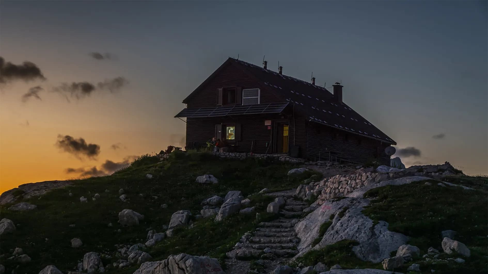 A wooden mountain chalet on a hill is bathed in warm light from a window. The sky, tinged with shades of orange and blue, indicates twilight. Rocks scatter