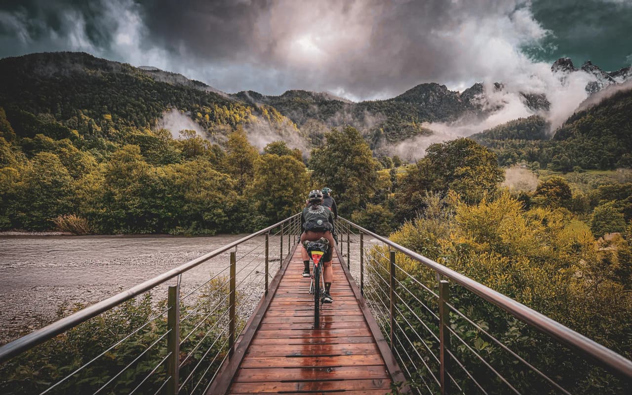 A cyclist crosses a wooden bridge, surrounded by lush greenery and majestic mountains. The sky is cloudy, adding a dramatic atmosphere to the scene. Mists float over the