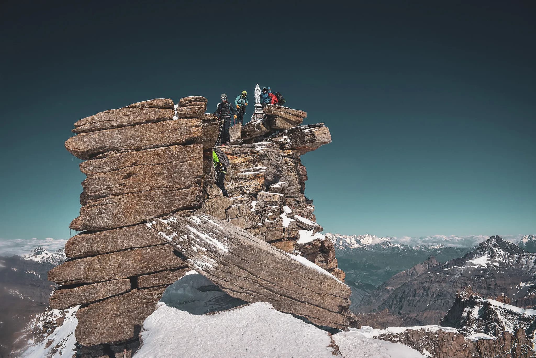 Alpinisten Gran Paradiso beklimmen via een smalle, rotsachtige bergkam. Enkele klimmers zekeren zich met touwen terwijl ze de top naderen. Op de achtergrond besneeuwde bergtoppen en een weids uitzicht.