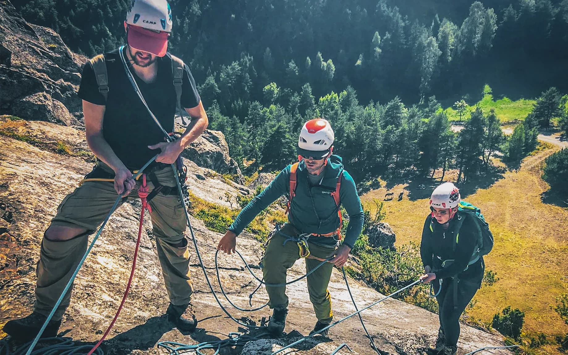 Three people climbing a cliff, wearing safety helmets and harnesses. One of them is managing the ropes, while the others move forward, taking care with their holds.