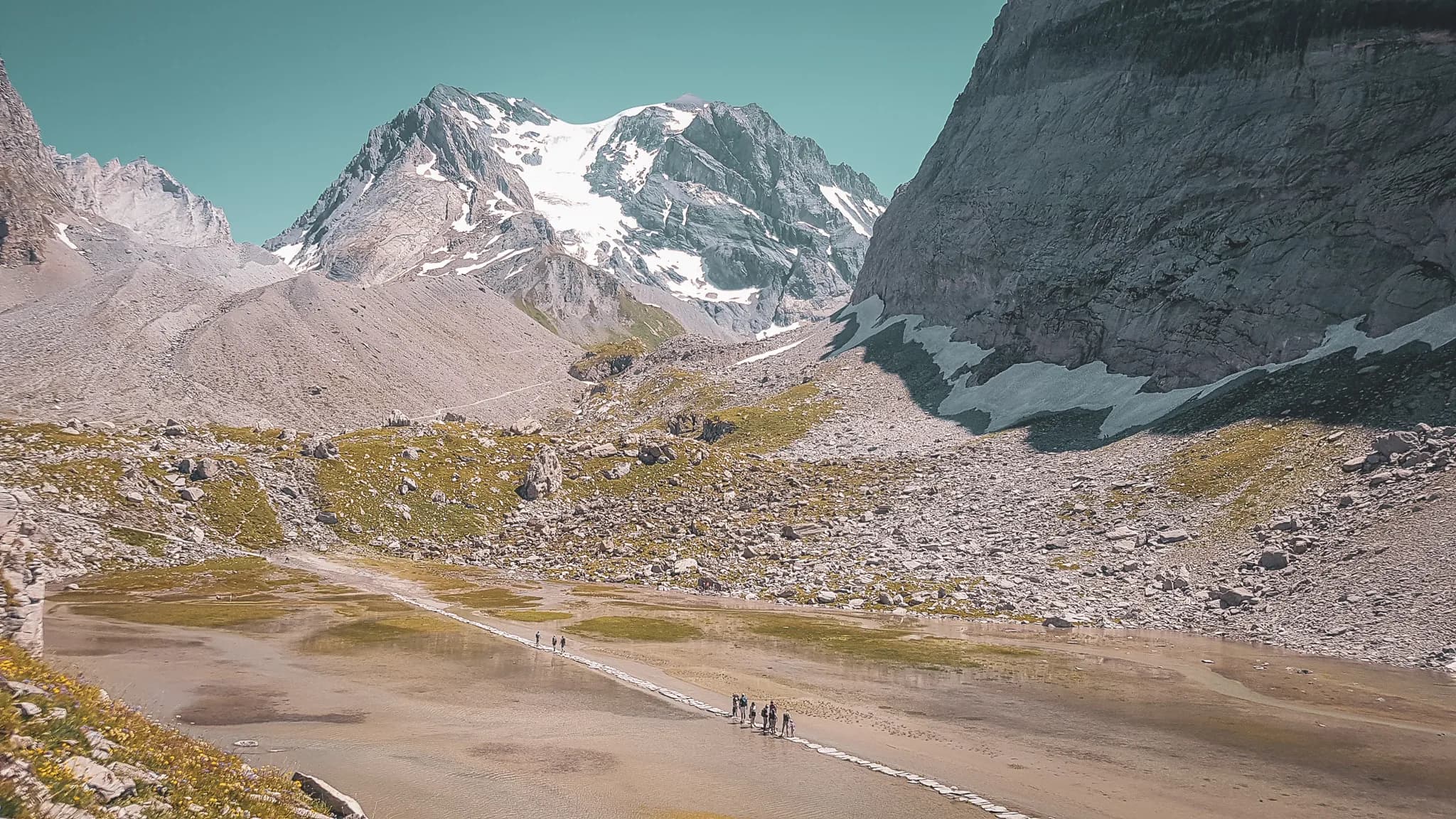 Un chalet en pierre est situé au pied de majestueuses montagnes, entouré de verdure. La construction présente un toit en tuiles et des balcons en bois, offrant une ambiance rustique et accueillante. Des arbres et des bu