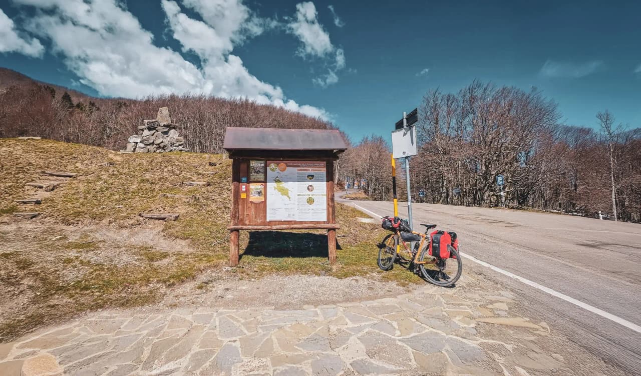 A wooden information board stands on slightly sloping ground, surrounded by green meadows and a few bare trees in the background. Next to the sign, a bicycle with panniers is parked.