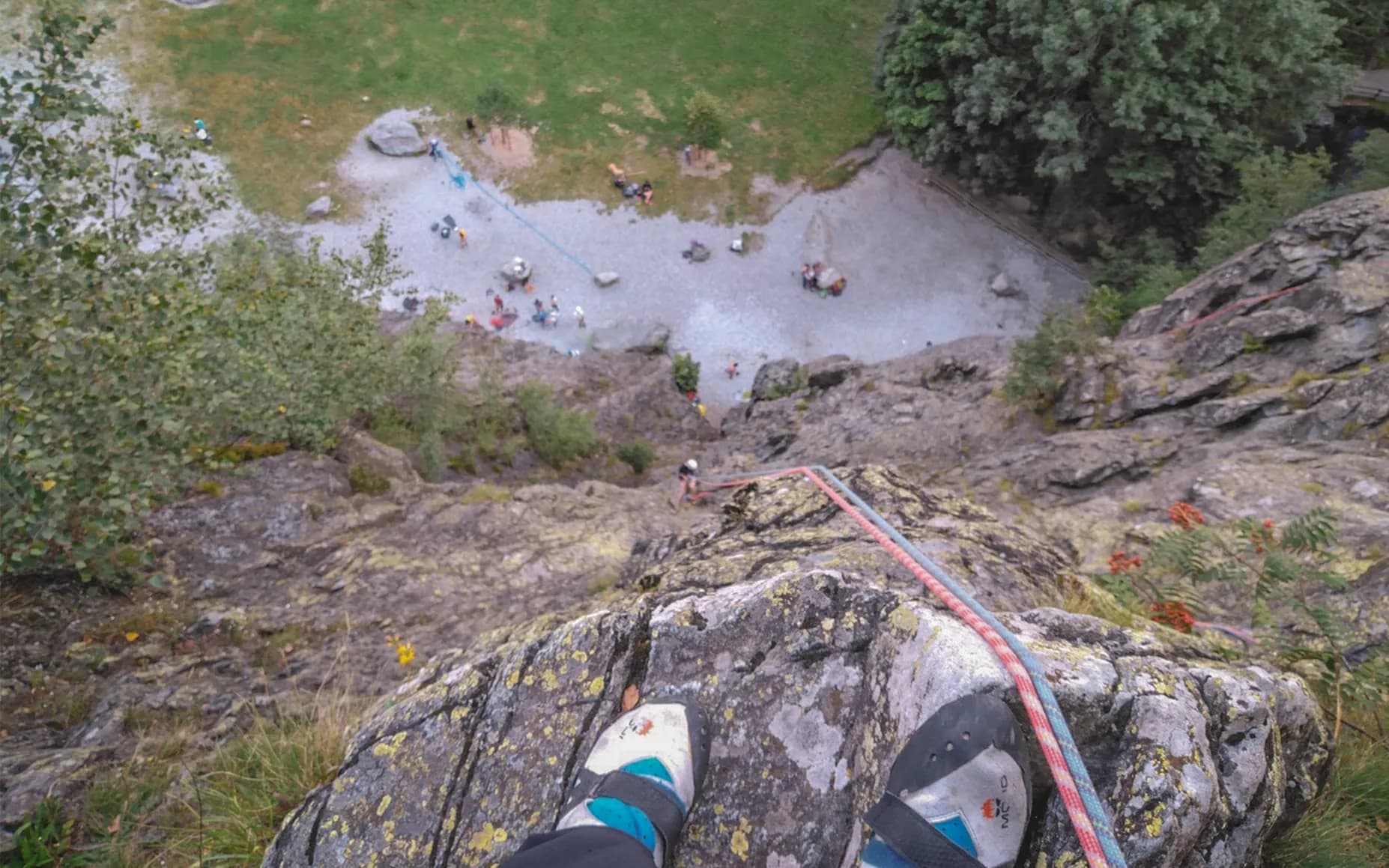 A bird's eye view from the top of a cliff, showing climbers and people on the ground below. The ground is gravel and grass, with a few rocks scattered about. Nearby