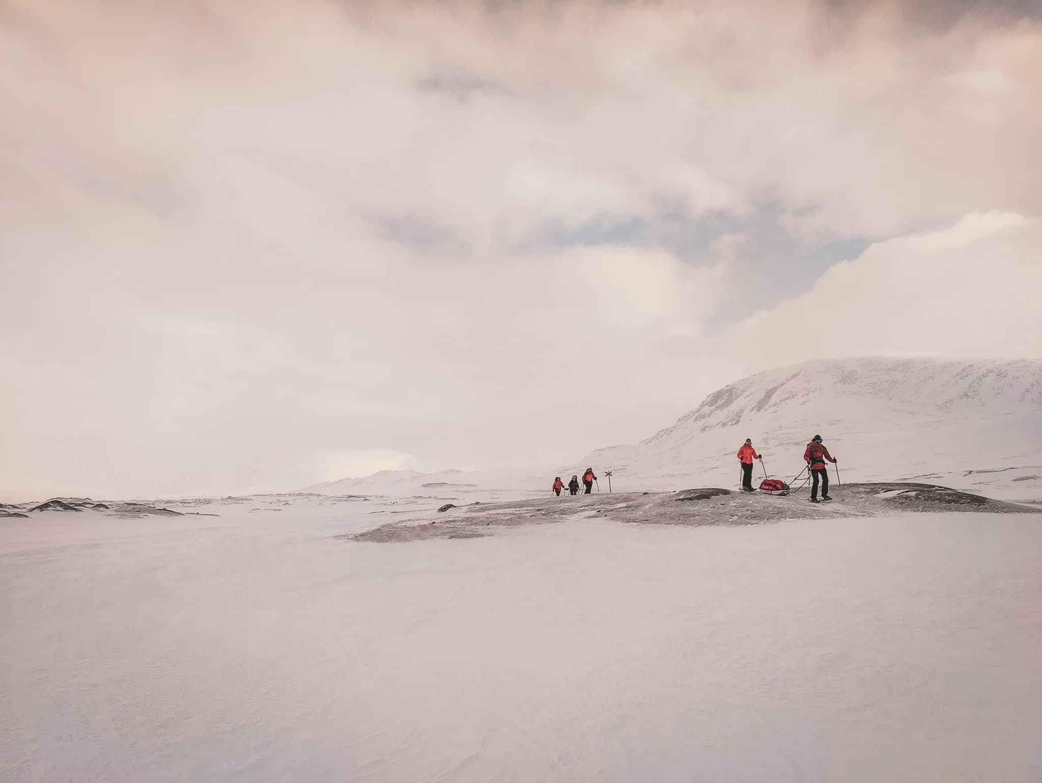 A winter landscape reminiscent of a wilderness adventure, with several hikers in red outfits making their way across snow-covered terrain. The hikers, equipped with pulkas, move slowly on snowshoes.