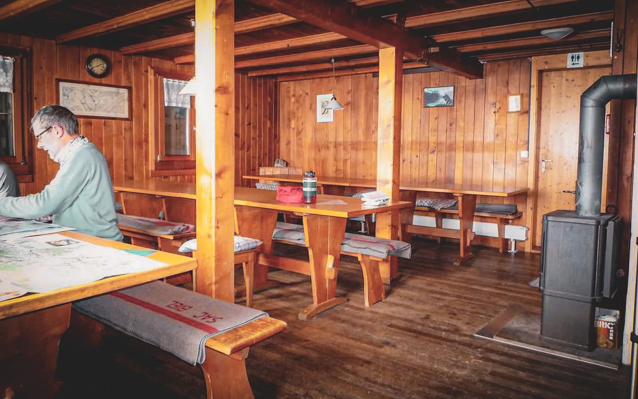 The interior is decorated in wood, with wood-panelled walls and ceiling. Two solid wood tables are in the centre, with cushioned benches. On one of the tables, several cards are spread out.