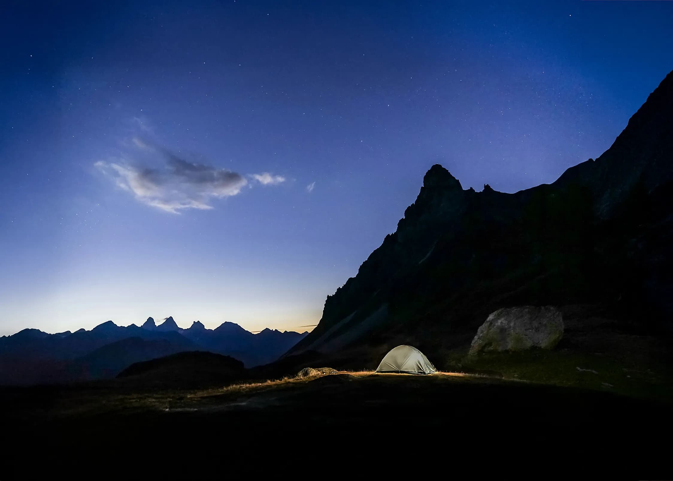 A mountain landscape at dusk, with a lighted tent in the foreground. In the background, mountain peaks stand out against a starry sky, tinged with blue and violet, while a