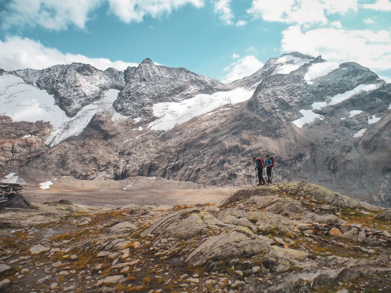 Twee wandelaars bewonderen de majestueuze bergen en gletsjers onder een blauwe hemel.
