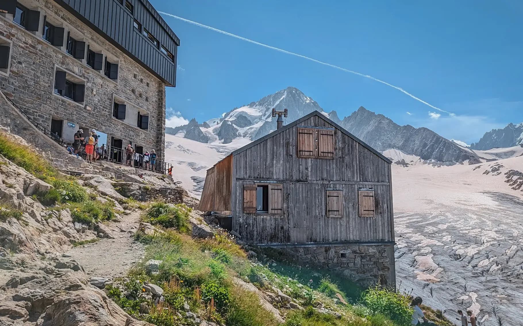 A stone and wood building is set in a mountainous landscape, surrounded by rocky heights and glaciers. Nearby, tourists gather on a stone path, admiring the view. The sky is blue