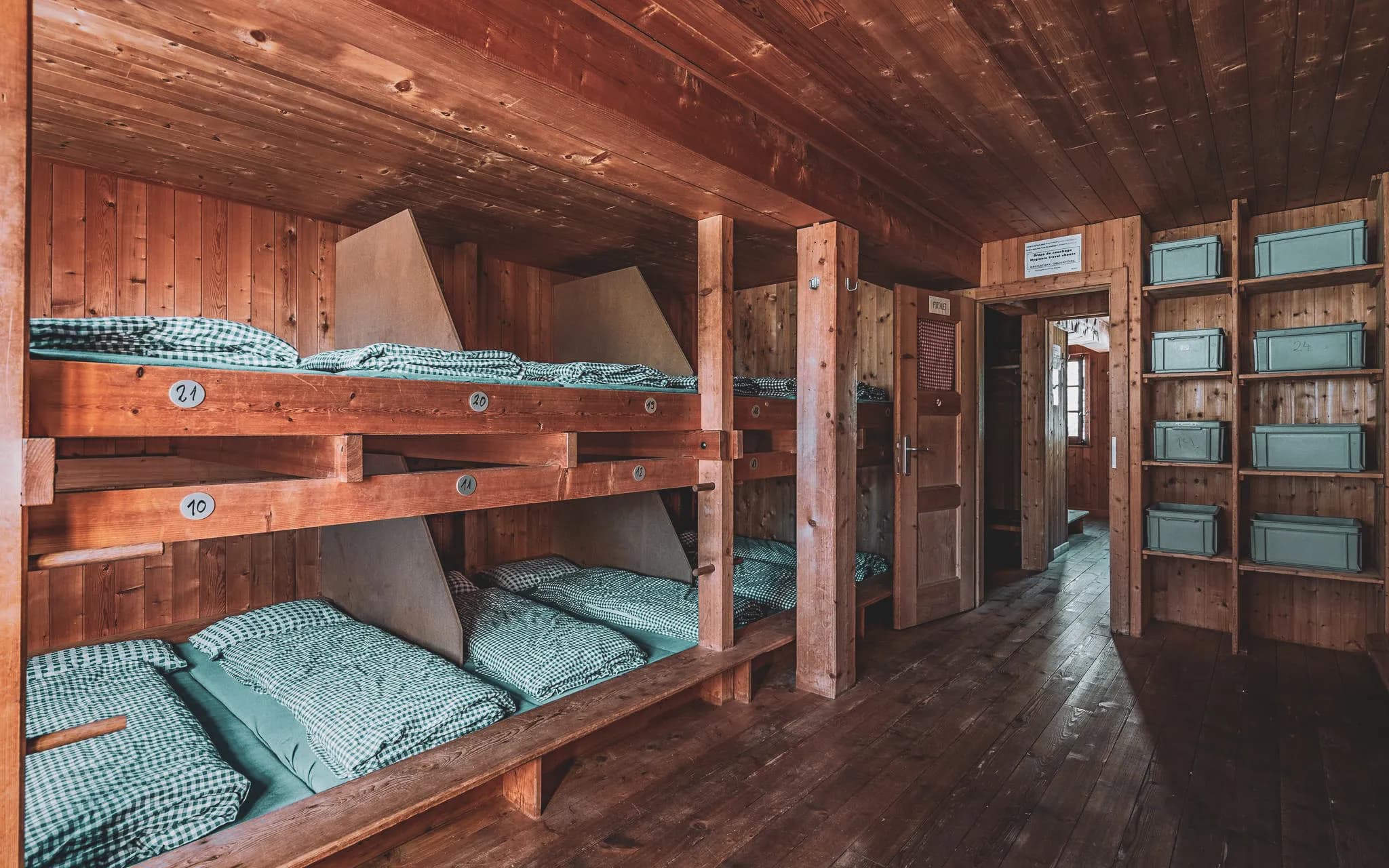 A shared room, typical of a wooden refuge, with wooden bunk beds. Each bed is furnished with green and white checked blankets, numbered 10 to 21.