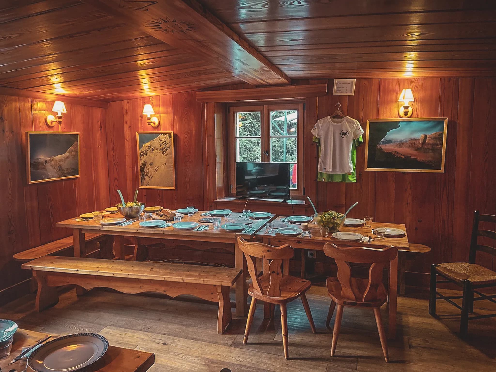 The warm ambience of a wooden interior, with a large wooden table decorated with crockery. Wooden chairs surround the table, while wall lamps create a soft glow. Two images