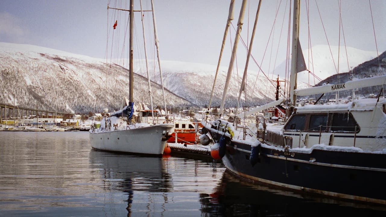 Two sailing boats moored in the harbour at Tromsø, surrounded by snow-capped mountains and a clear sky.