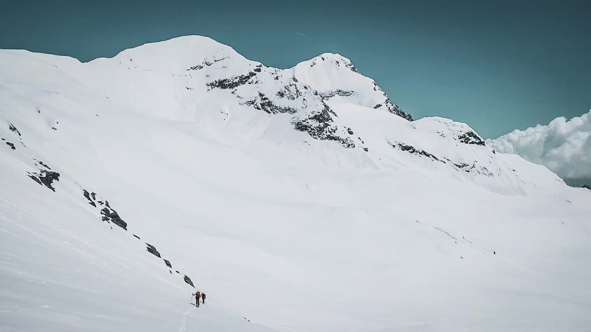 A snow-covered mountain landscape, with imposing snow-capped mountains in the background. In the foreground, two hikers are making slow progress over an icy surface, creating tracks in the snow.