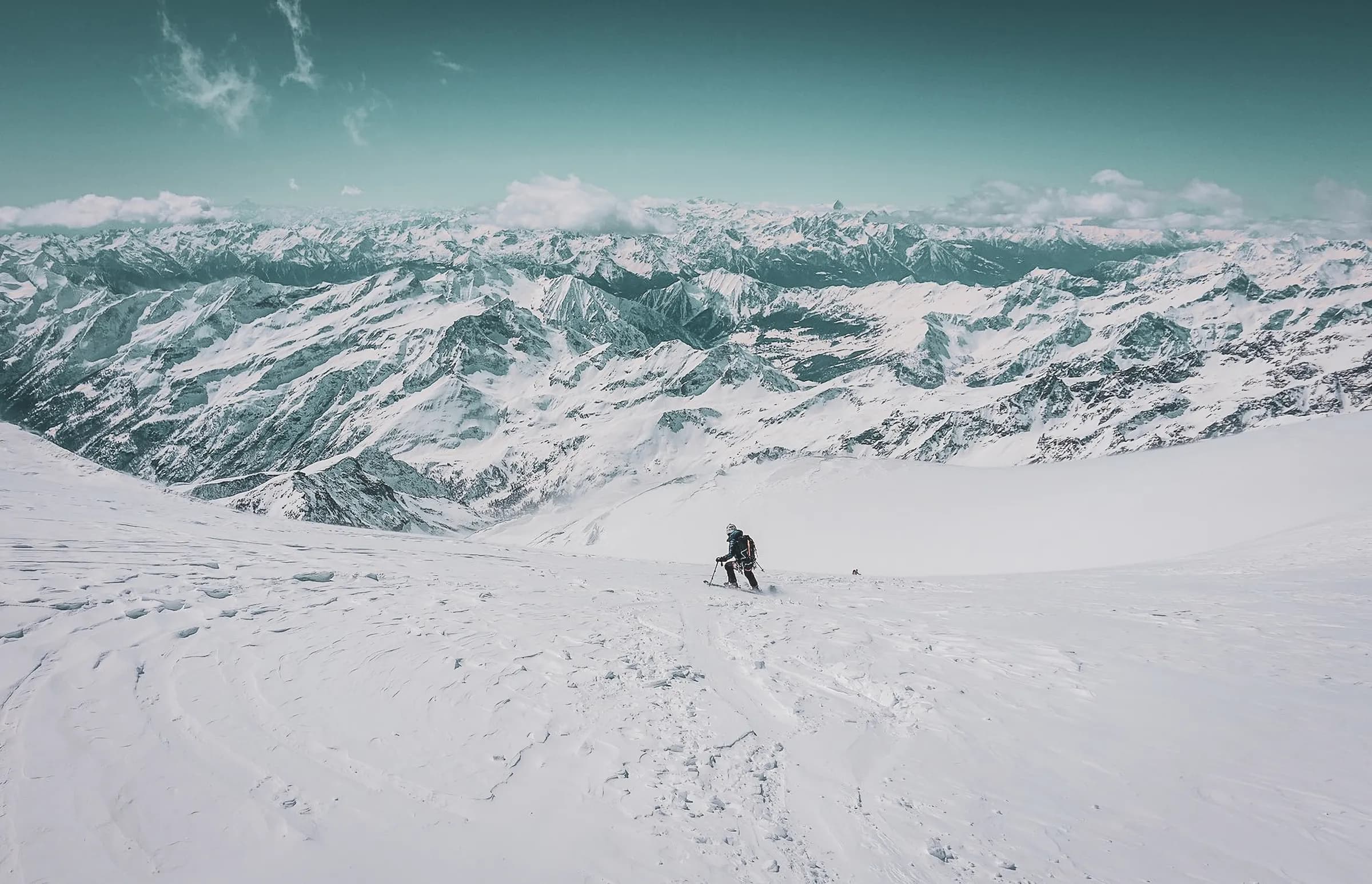A snow-covered mountain landscape stretches as far as the eye can see, with imposing peaks covered in snow. A mountaineer, equipped with hiking gear, makes his way through the snow. The sky has a