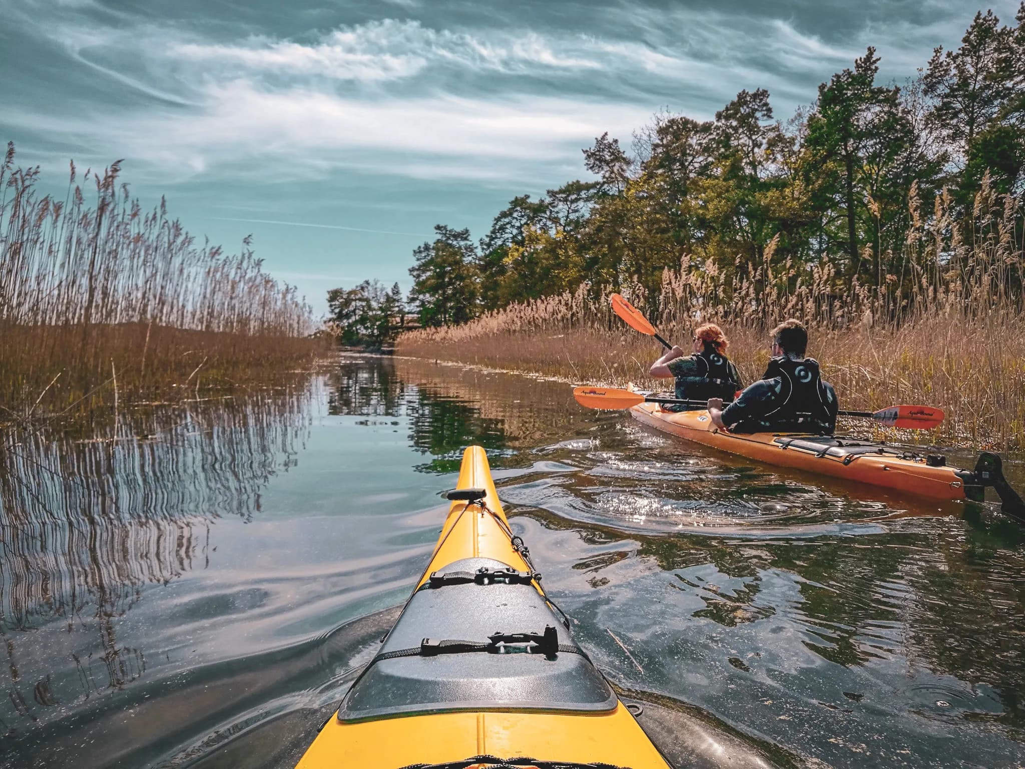 A peaceful waterscape with a view from a yellow kayak towards two orange kayaks. The kayaks glide over calm water, surrounded by tall dry grass and tree foliage on