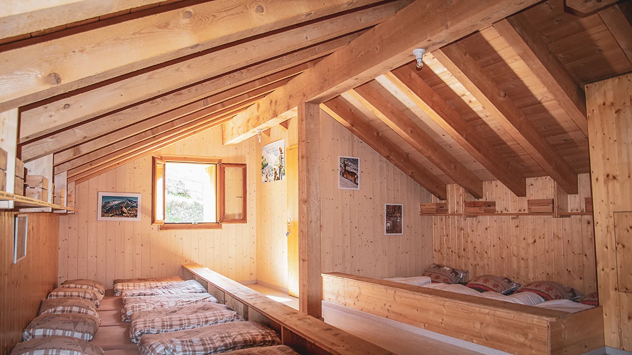 A rustic-style bedroom with wooden floors and walls and a sloping ceiling. To the left, several mattresses stacked on a wooden platform. On the right, a large double bed with decorative pillows.