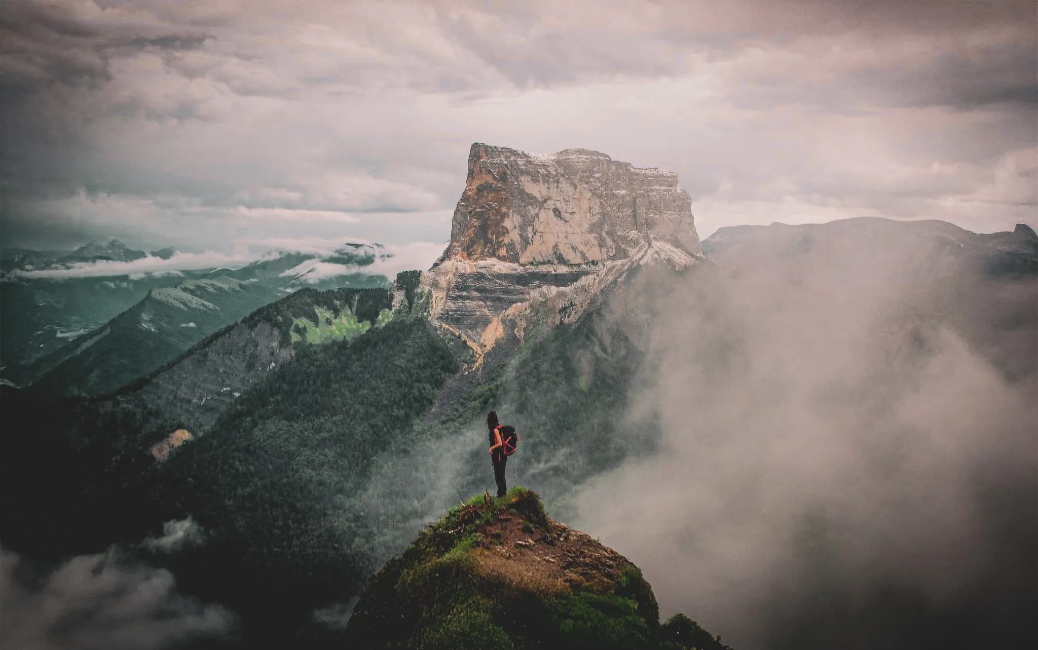 An adventurer stands on a rocky promontory overlooking a majestic landscape of mountains and valleys. The sky is cloudy, creating a dramatic atmosphere, and fog banks