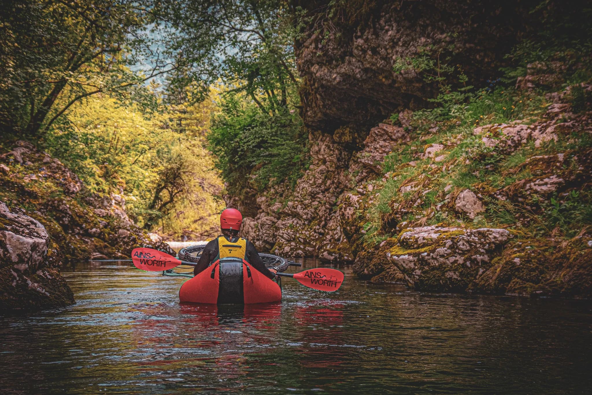 A kayaker navigates through a narrow waterway lined with rocks and lush vegetation. The water is calm and reflects the surrounding scenery, creating a peaceful atmosphere. The kayaker,