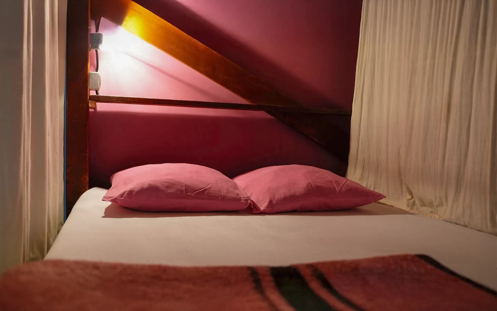 A single bed with two pink pillows, set in a room with a pink wall. Soft light illuminates the space, creating a warm ambience. In the background, light fabric curtains frame the bed, complementing the pink wall.