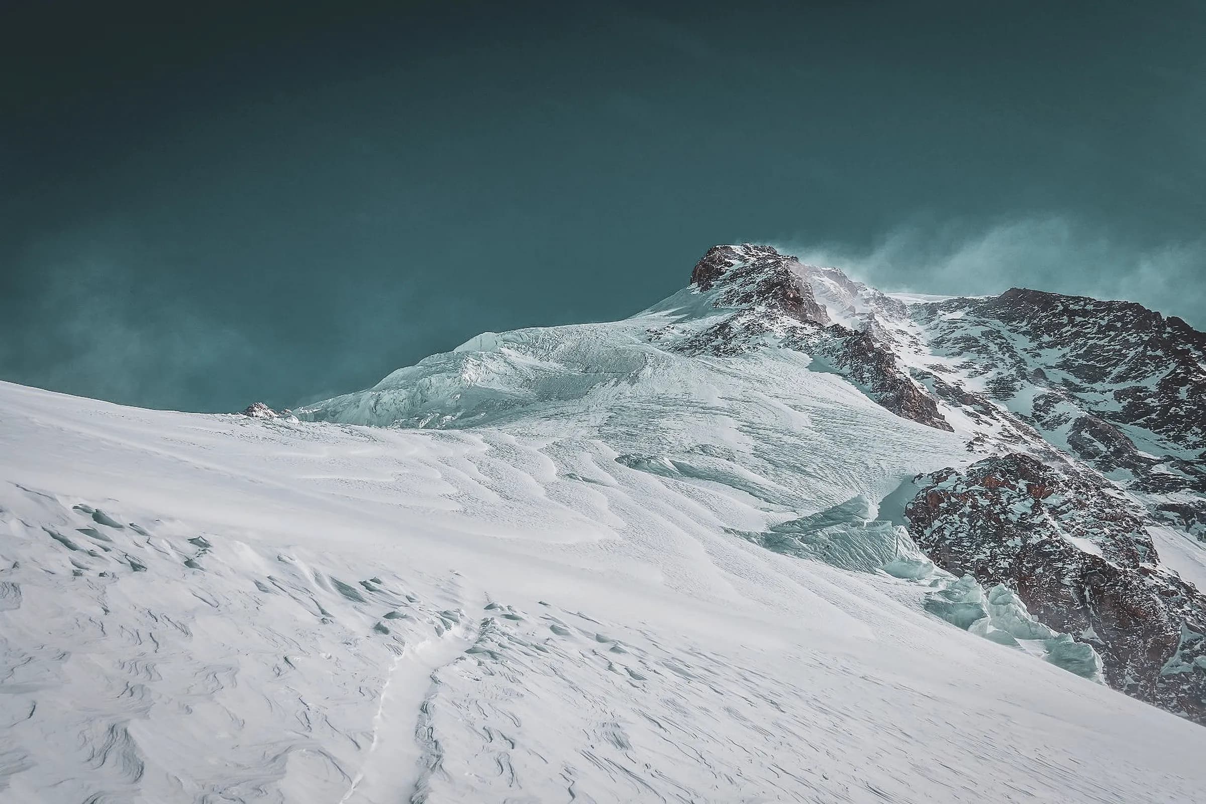 A majestic mountain peak covered in snow, with glacial slopes sculpted by the wind. The clear, deep blue sky contrasts with the brilliant whiteness of the snow. Rock formations