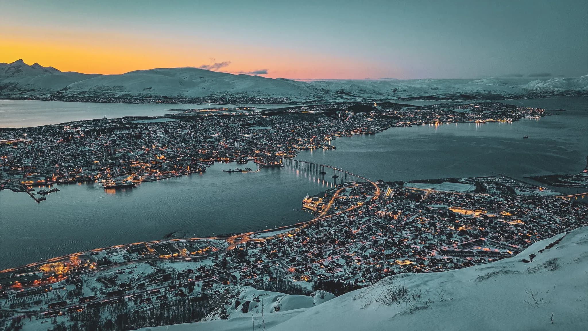 Panoramic view of the snow-covered town of Tromsø, lit up at dusk and surrounded by majestic fjords.