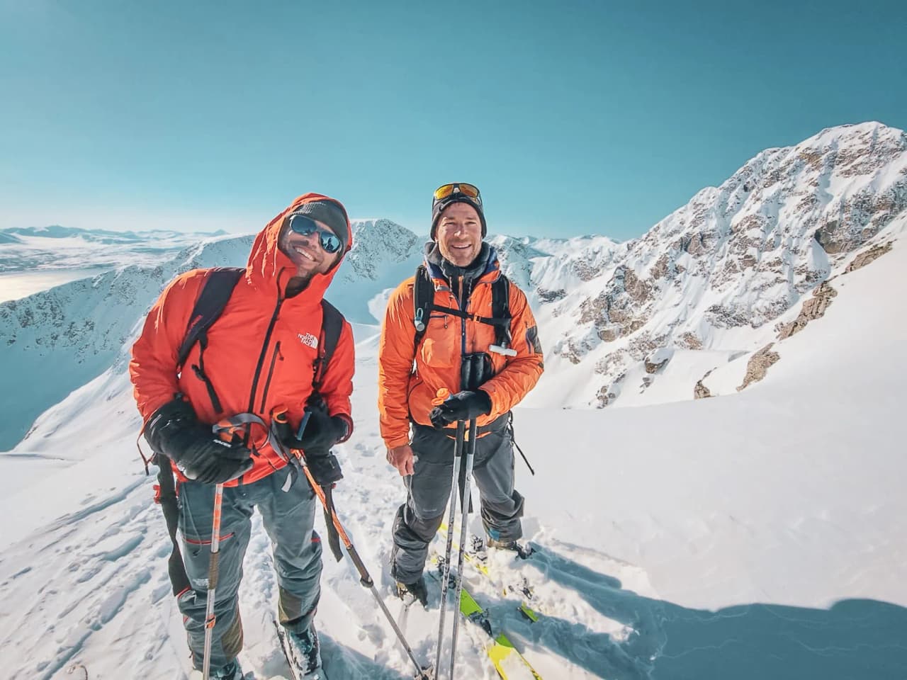 Two smiling climbers stand on a snow-covered summit. They are wearing orange jackets, protected for the cold conditions, and are surrounded by majestic mountain scenery under a clear blue sky. One of them is