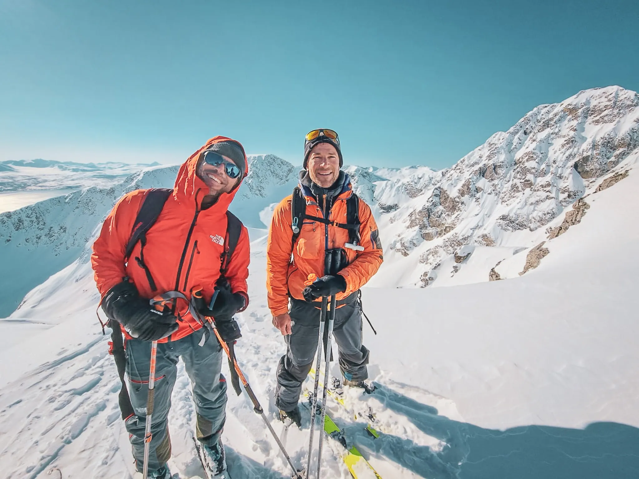 Two smiling climbers stand on a snow-covered summit. They are wearing orange jackets, protected for the cold conditions, and are surrounded by majestic mountain scenery under a clear blue sky. One of them is