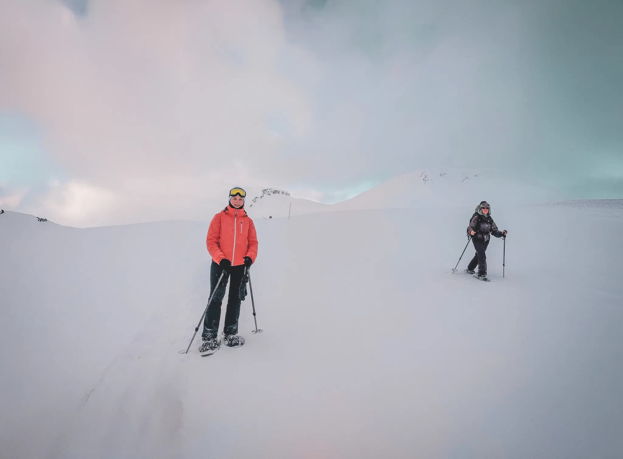 A snowy landscape with two people in ski gear. One, wearing an orange coat and ski goggles, is standing on the snow, while the other is dressed in black and wearing a balaclava.
