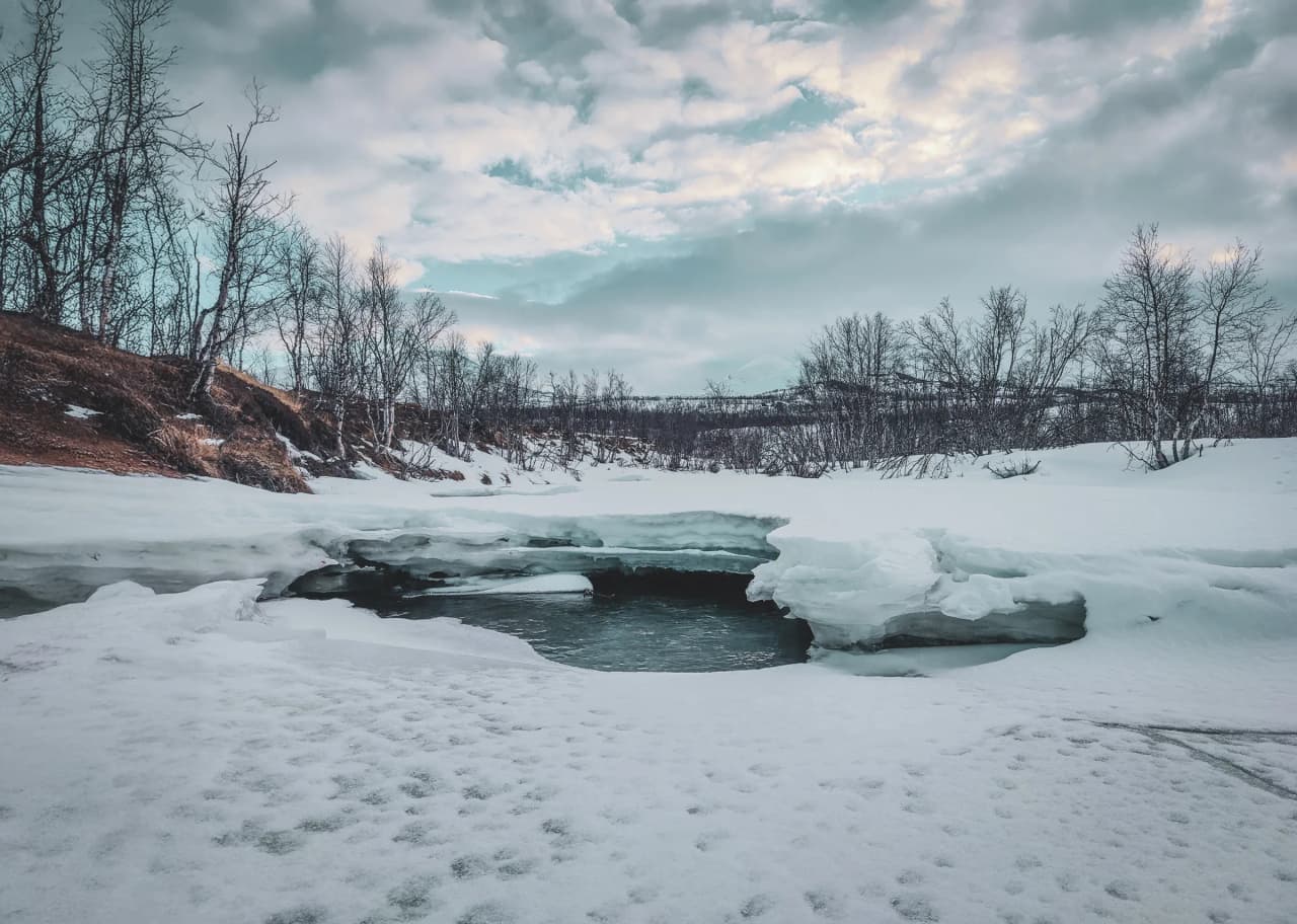 A captivating winter landscape, with snow-covered hills, bare trees and a partially frozen stream.