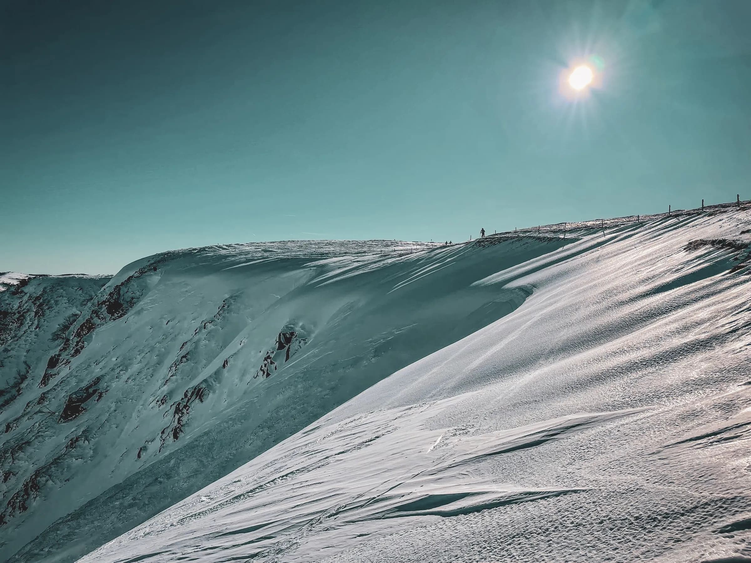 Een uitgestrekt besneeuwd landschap met glooiende hellingen en een scherp reliëf. De sneeuw glinstert in de felle zon en creëert een serene, rustgevende sfeer. Aan de horizon, een lijn