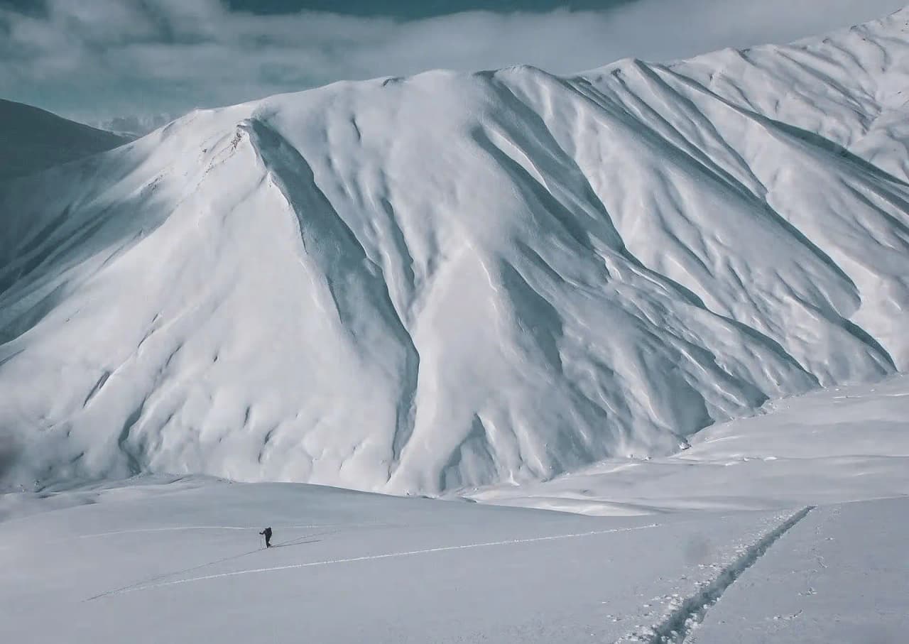Un skieur solitaire trace des sillons sur une neige immaculée, face à des montagnes majestueuses.