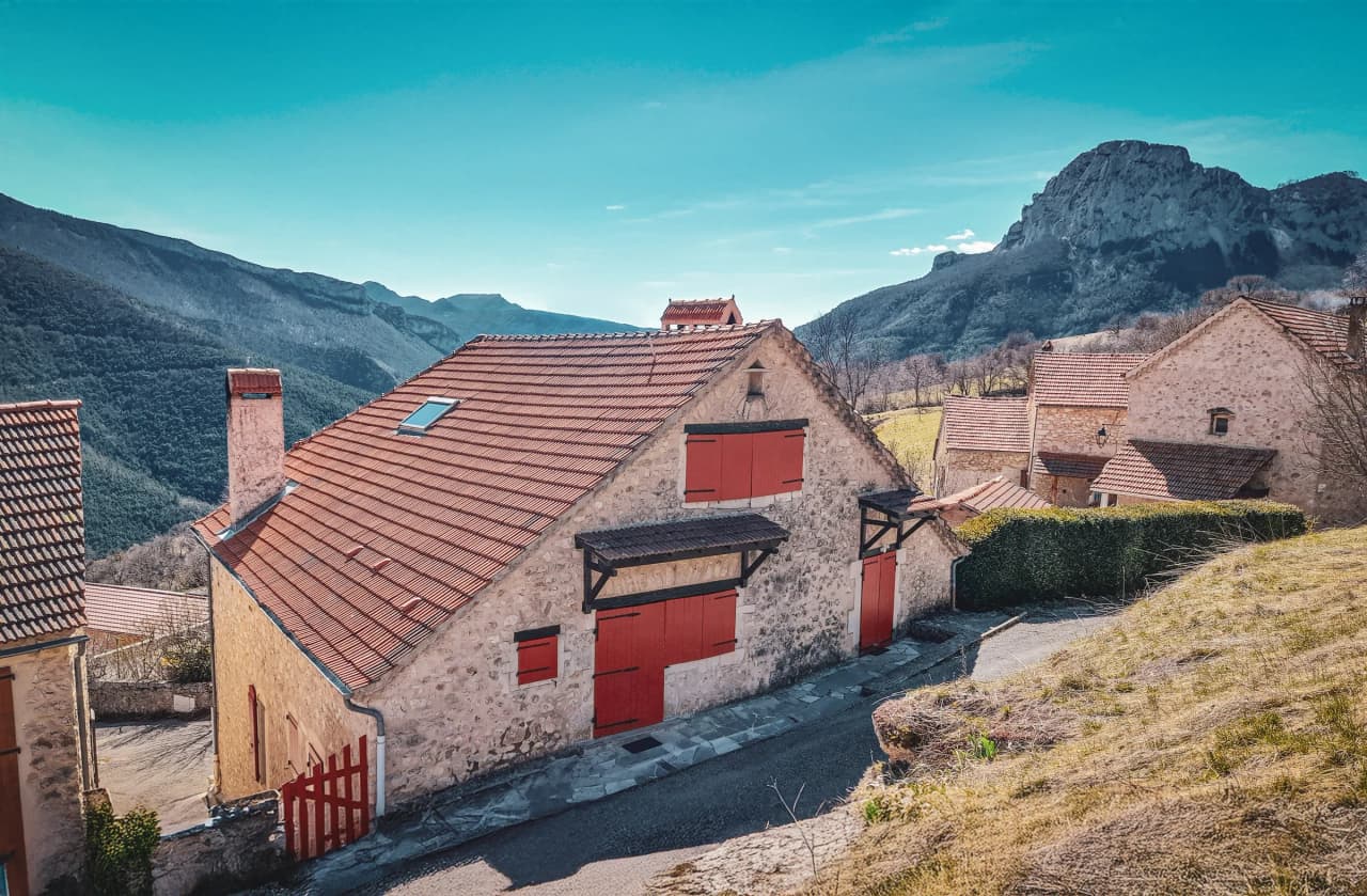 Charmante maison en pierre avec volets rouges, entourée de montagnes verdoyantes sous un ciel bleu.