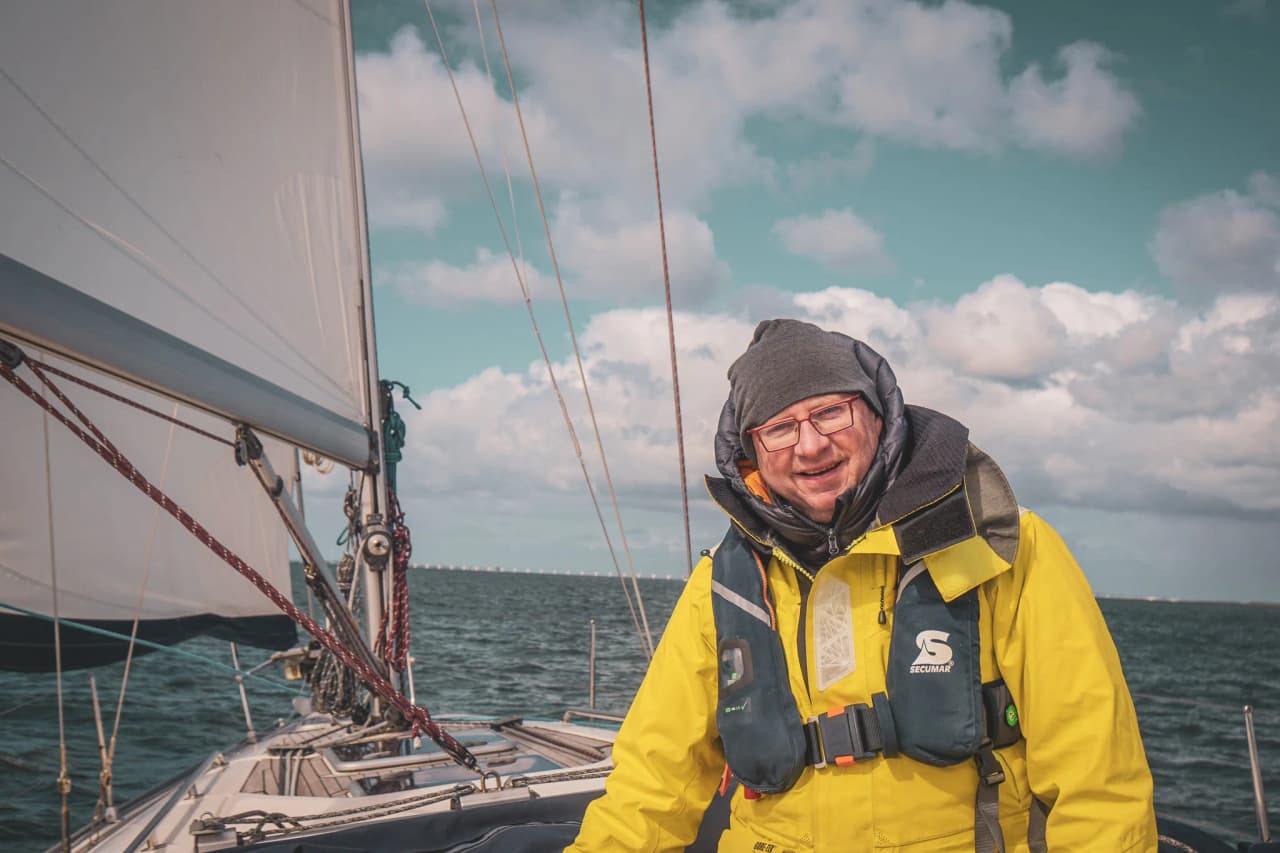 Een lachende man in een geel wetsuit op een zeilboot, varend door rustige wateren onder een bewolkte hemel.