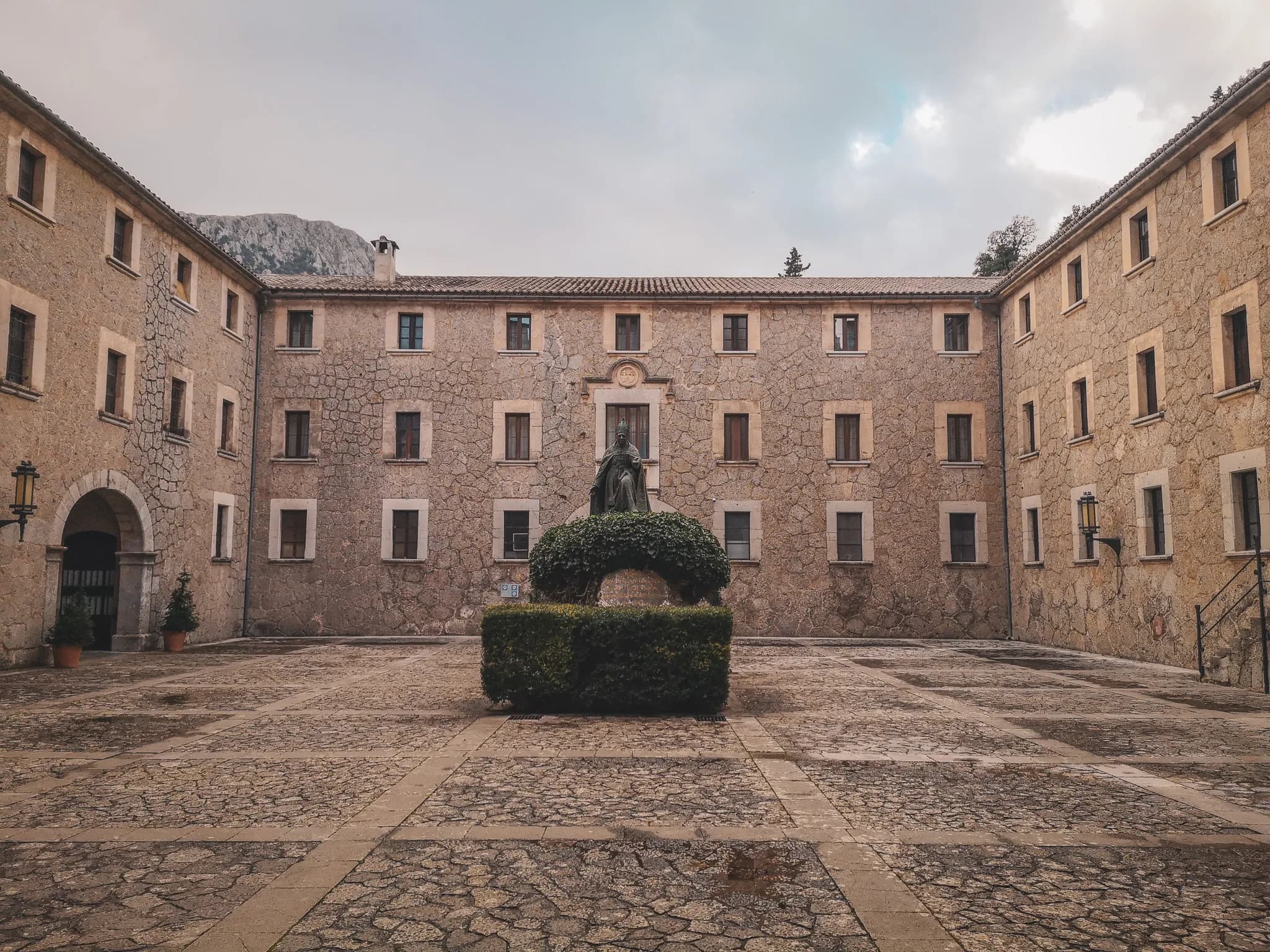 A stone courtyard surrounded by stone buildings, with a verdant statue in the centre, inviting you to explore.