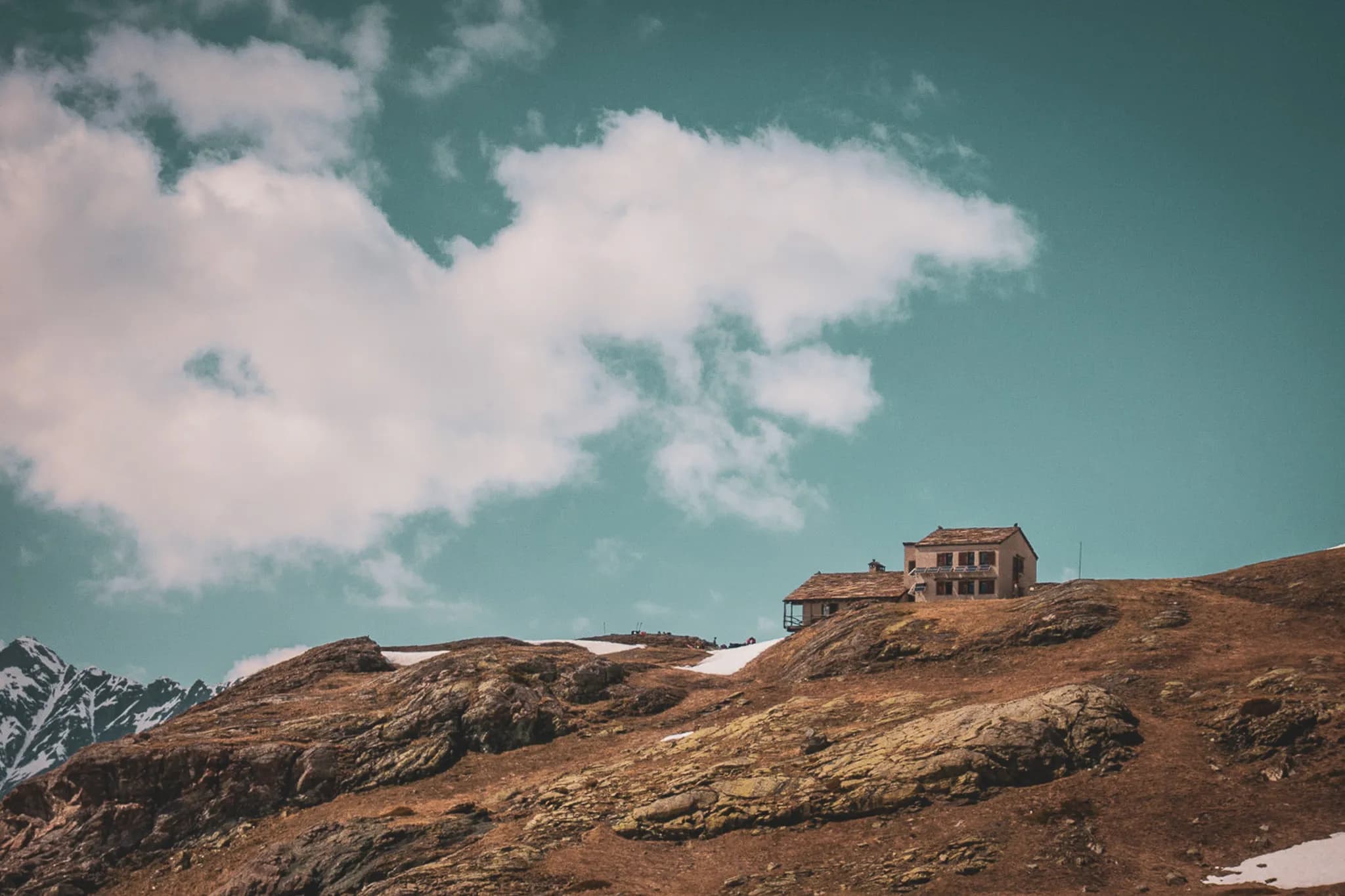 An authentic chalet stands on a rocky hill, surrounded by mountain scenery. The beige and brown building has a tiled roof. In the background, the peaks of the