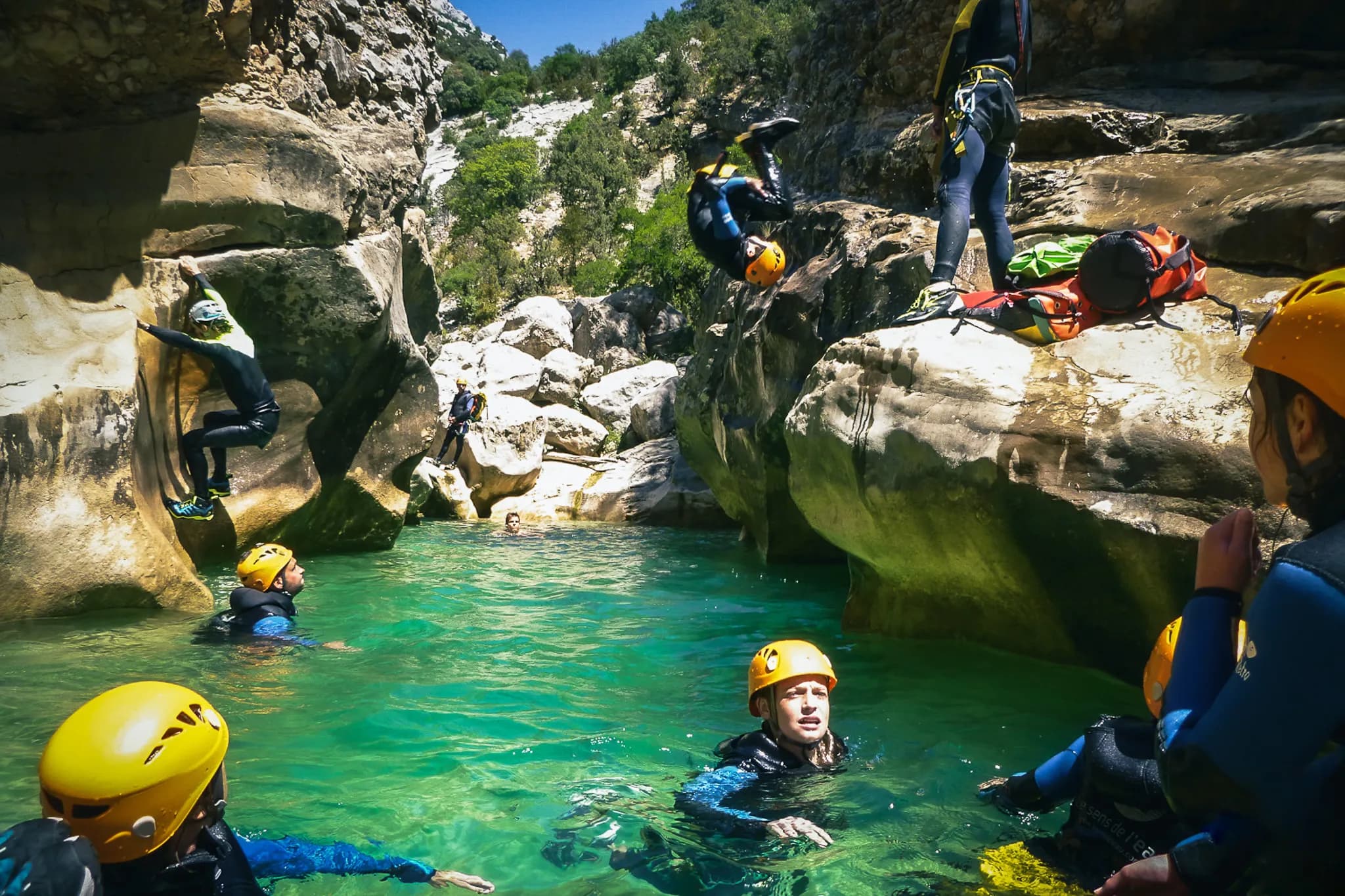 A group of people in wetsuits and helmets are in a natural environment, near a turquoise stream surrounded by rocks. Some of the participants are in the water, while others are climbing up rock faces.