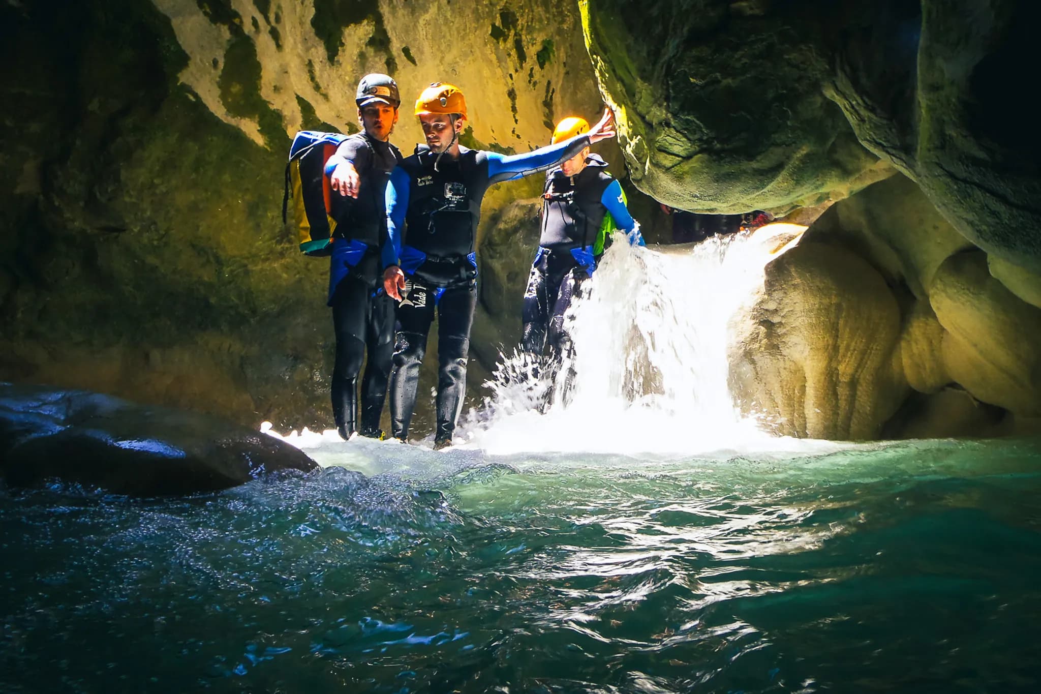 A group of people dressed in diving suits are in a clear river, surrounded by rock faces. One of the participants, wearing an orange safety helmet, places his hand on a stone vo