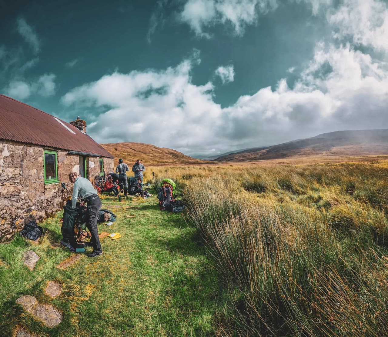 Een groep avonturiers bereidt zich voor op hun trektocht bij een stenen hut in het hart van de Schotse Hooglanden.
