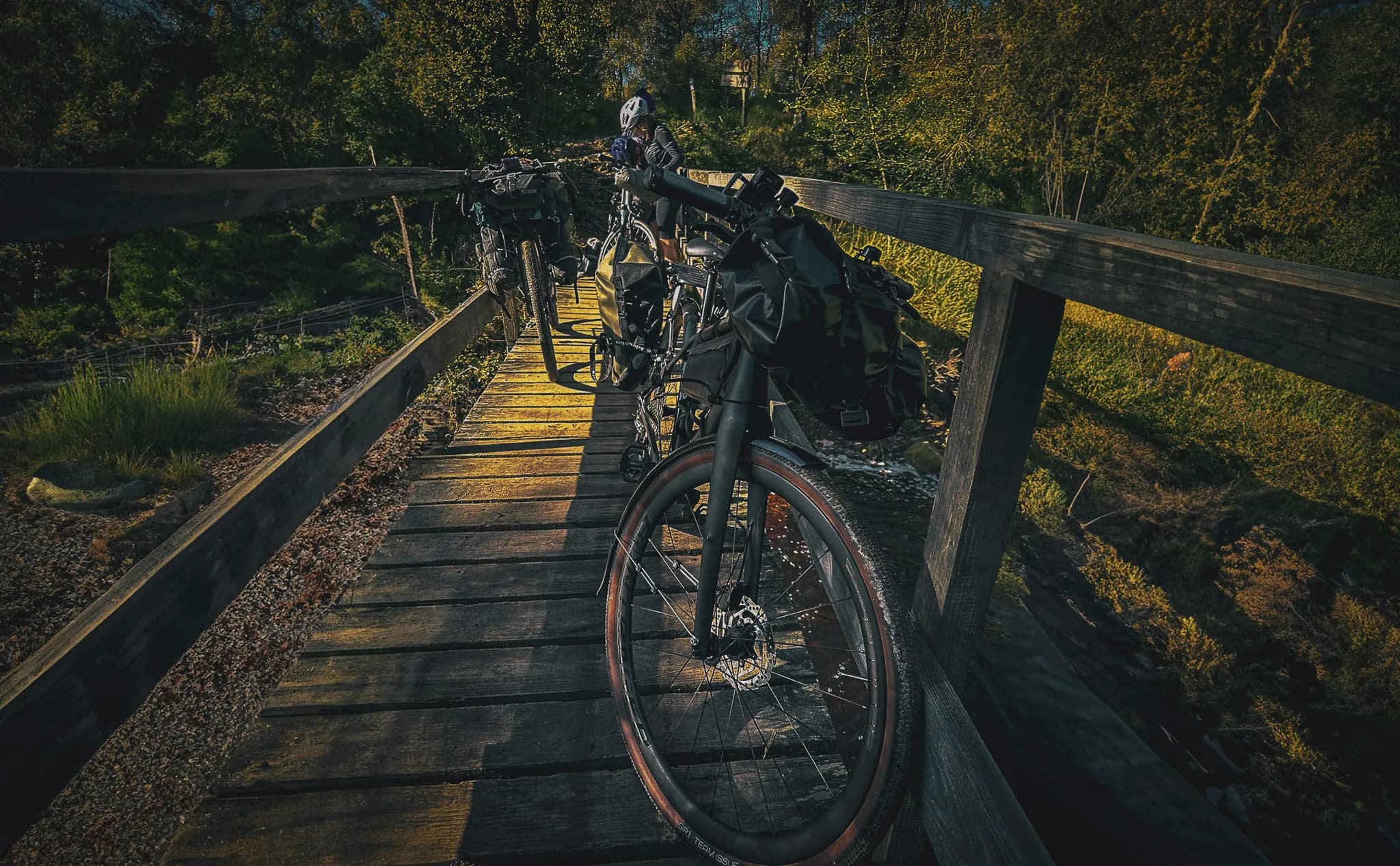 A wooden path overlooks a small stream, surrounded by lush greenery. Two bicycles loaded with travel bags are parked on the bridge, while one cyclist is checking his phone.