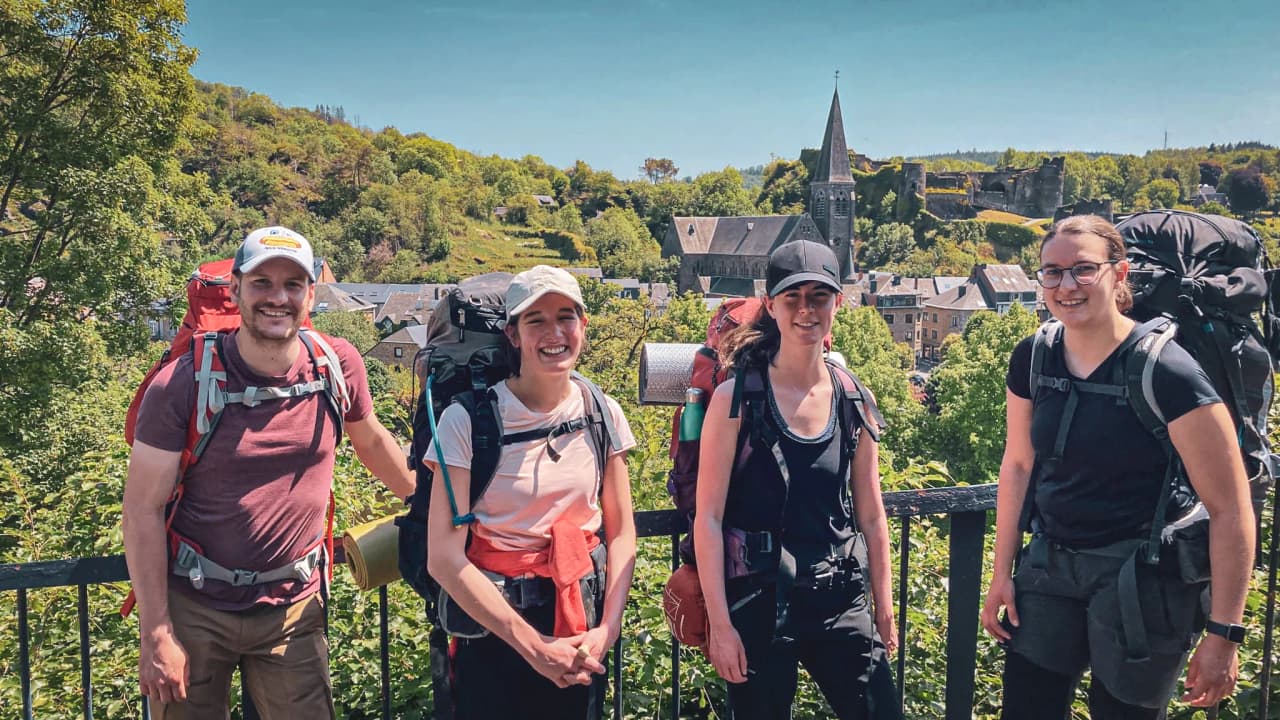 Quatre jeunes aventuriers souriants, sacs à dos prêts, devant un paysage verdoyant en Ardenne belge.