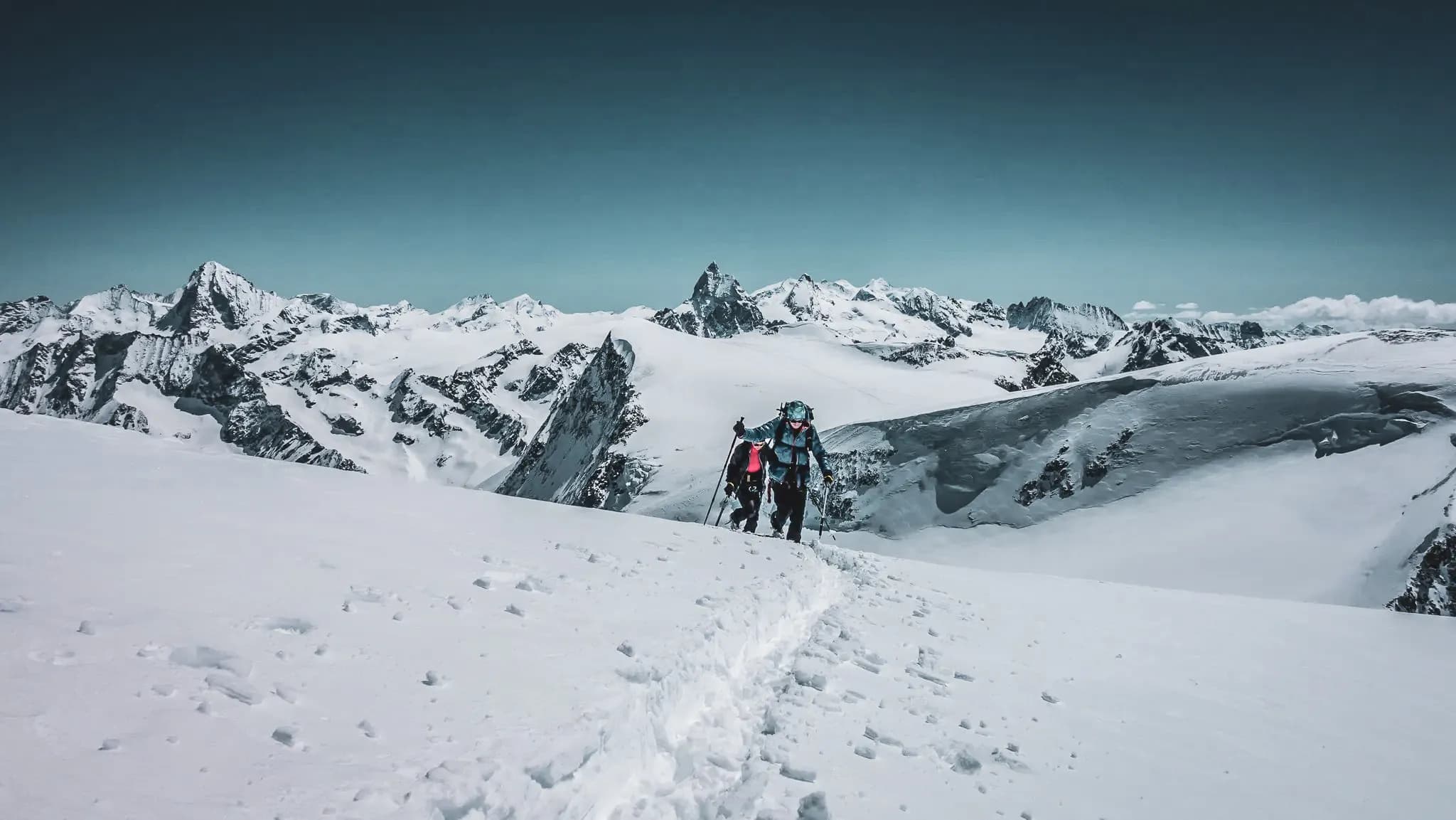A high mountain landscape covered in snow, with mountain bikers making their way along a path cut through the snow. The sky is clear, a soft shade of blue, and the majestic peaks of the