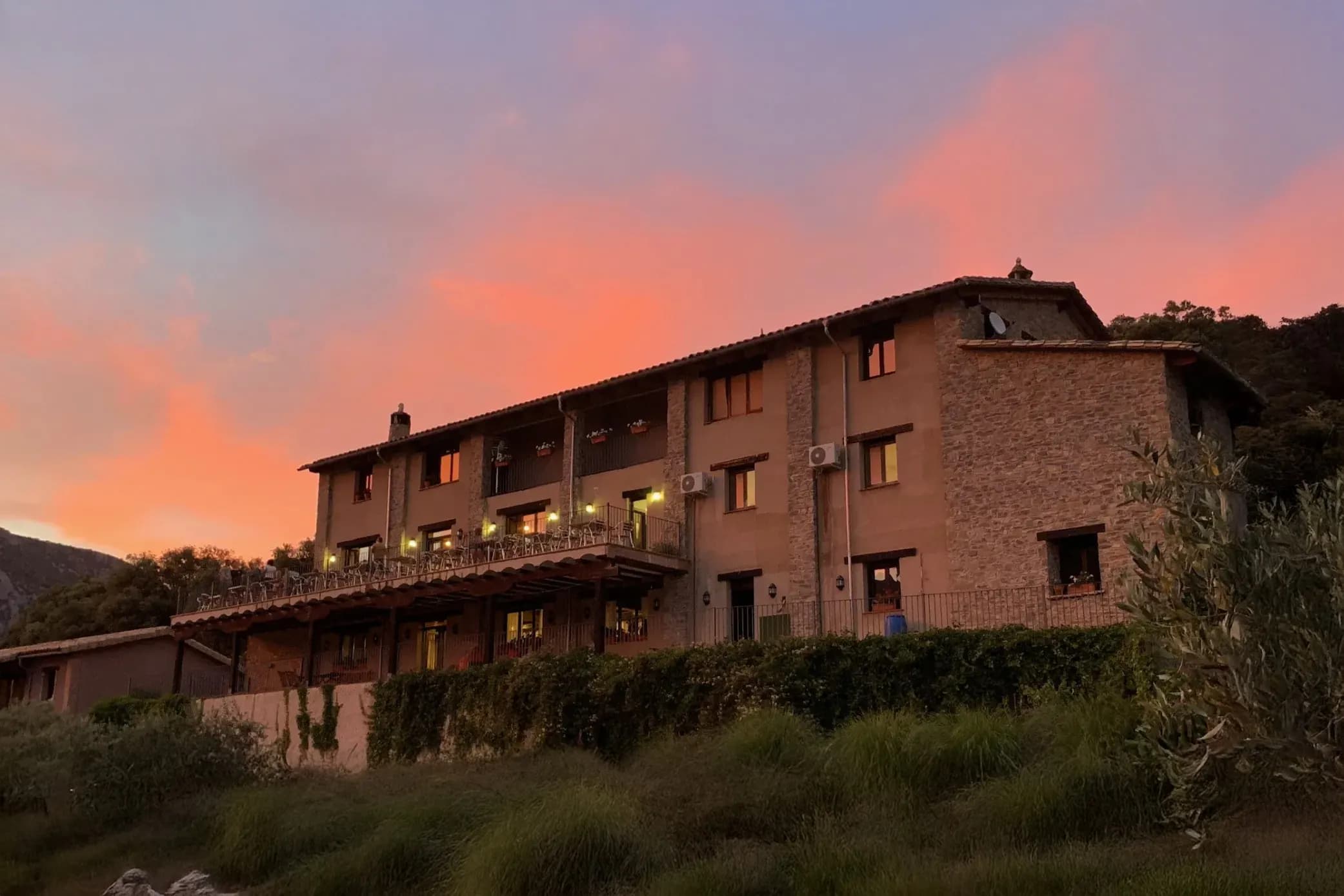 A stone building stands magnificently on a hill, with wooden balconies adorned with white chairs. The illuminated windows create a warm atmosphere at dusk, when the sky is tinged with