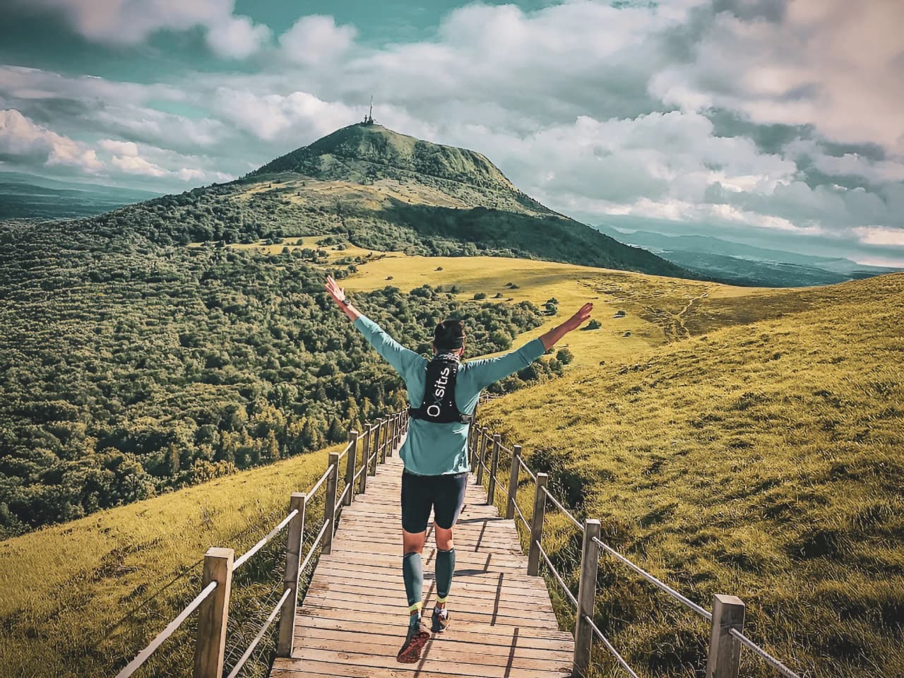 Un coureur sur un chemin en bois devant les volcans verdoyants d'Auvergne, en pleine nature.