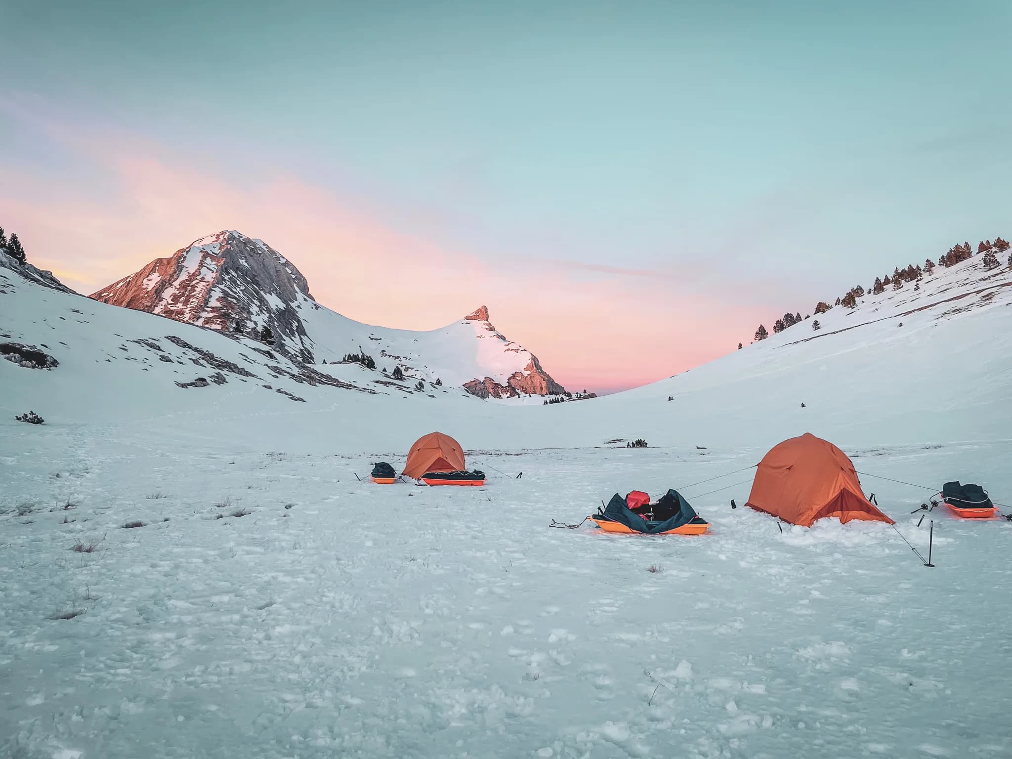 A snowy landscape with orange tents set in a vast field of snow. In the background, majestic mountains rise up, with peaks illuminated by the soft light of dusk, where the sky