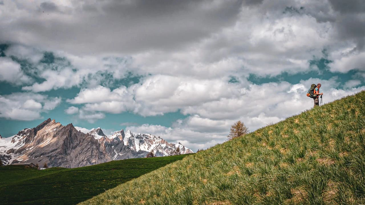 Un randonneur grimpe une colline verdoyante avec des montagnes majestueuses en arrière-plan.