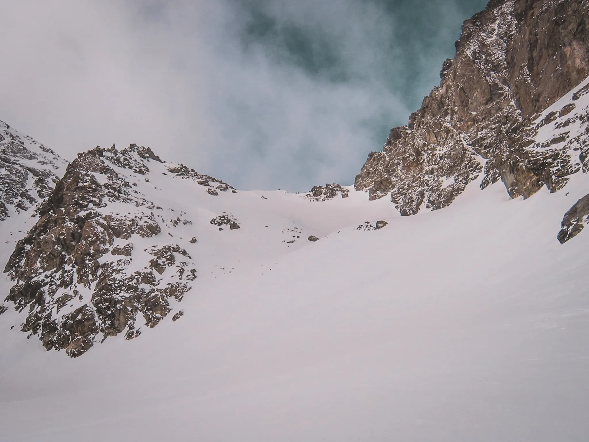 Majestic, snow-capped mountains rise up under cloudy skies. The landscape features steep slopes and rock formations, creating a stark contrast between the immaculate white of the mountains and the snow.