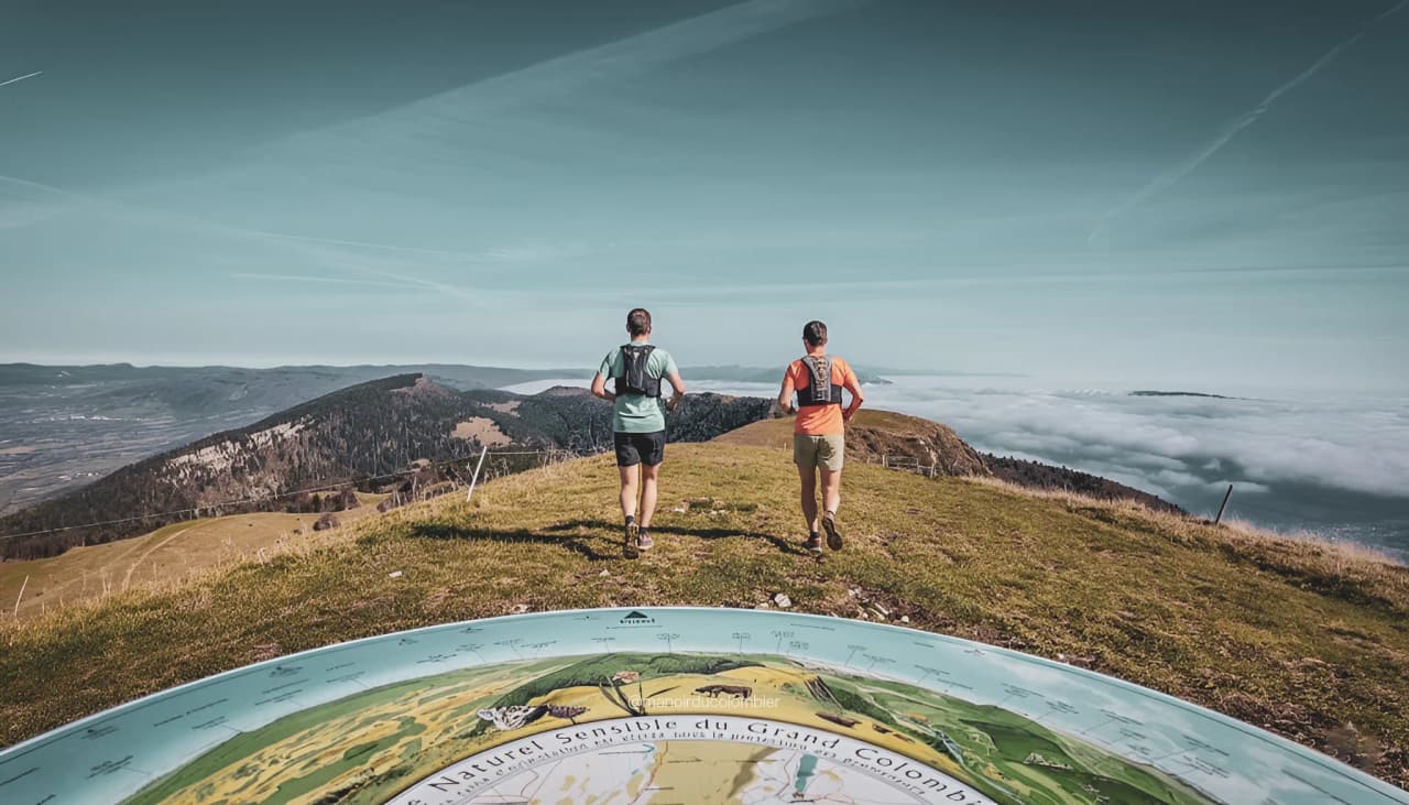 Two runners on a leafy trail at Grand Colombier, with a panoramic view of the mountains and clouds below.
