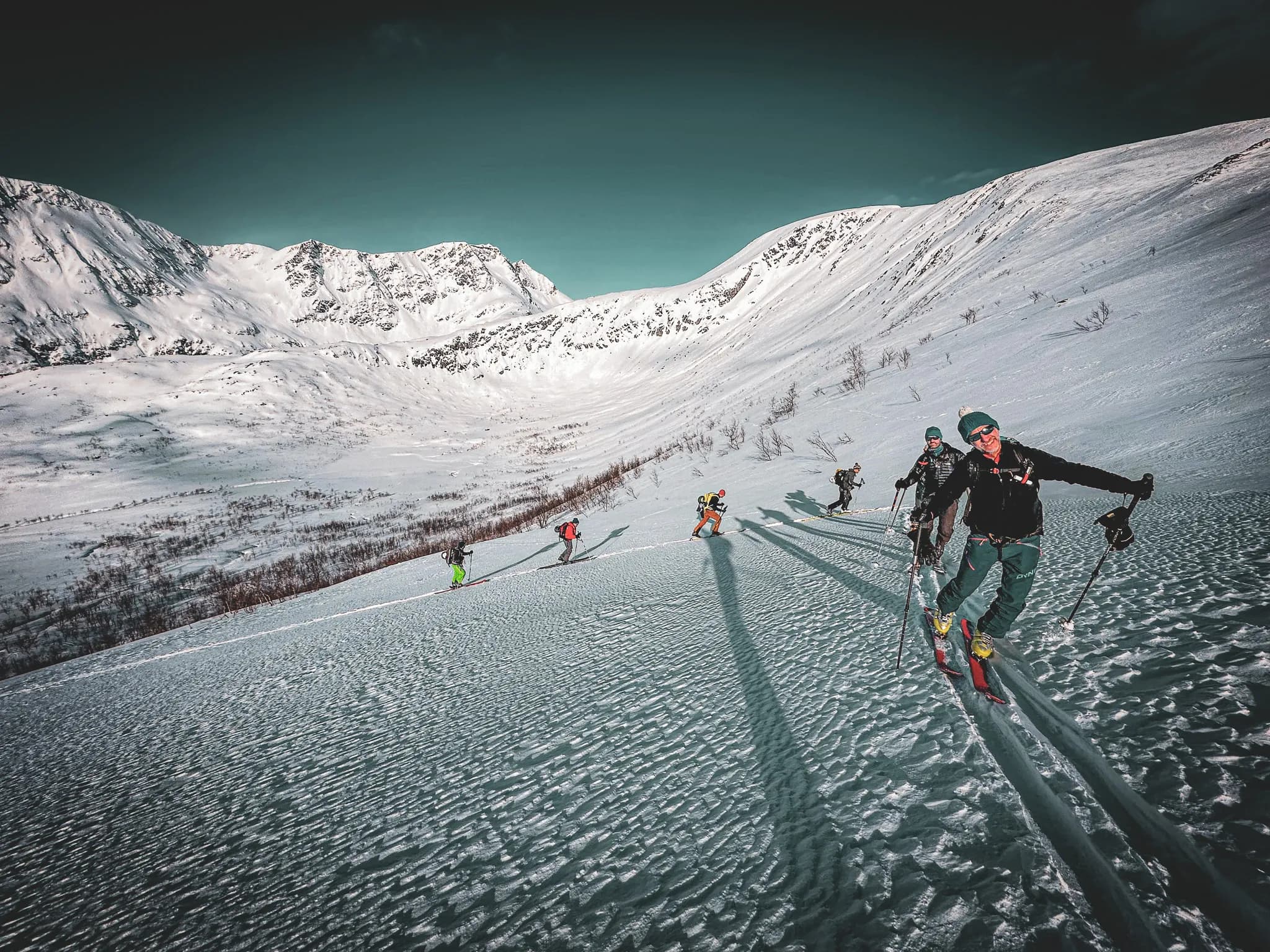 Un groupe de skieurs évolue sur une pente enneigée, entourés de montagnes majestueuses. Les skiants portent des équipements colorés, se déplaçant sur un manteau de neige fraîche, qui présente des motifs ondulés